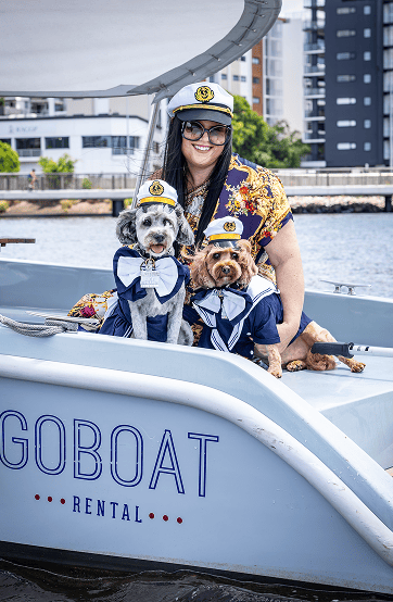 A woman wearing a captain's hat sitting in a boat with two dogs dressed in sailor outfits