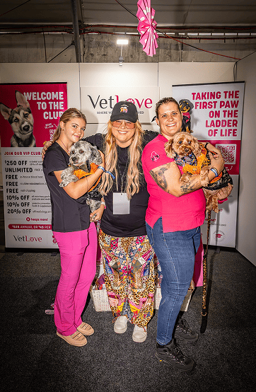 Three women holding two dogs at a VetLove branded promotional event backdrop