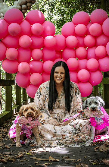 A woman sitting with two dogs in front of a giant heart-shaped pink balloon setup